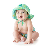 A baby smiles, wearing a Bumkins Sun Hat: Green Dinosaurs and a matching diaper with dinosaur patterns. The lightweight fabric offers UPF 50+ protection, while an adjustable drawstring ensures the hat fits snugly against a white background for playful comfort.