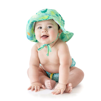 A baby smiles, wearing a Bumkins Sun Hat: Green Dinosaurs and a matching diaper with dinosaur patterns. The lightweight fabric offers UPF 50+ protection, while an adjustable drawstring ensures the hat fits snugly against a white background for playful comfort.
