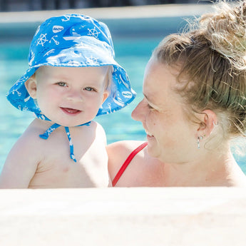 A woman in a red swimsuit holds a smiling baby wearing Bumkins Ahoy Sun Hat by the pool.