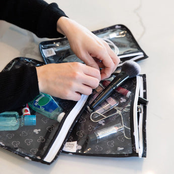 Hands organizing cosmetics in a TSA-compliant Bumkins Clear Bag featuring a Mickey Mouse Icon Black + White design on a white surface.