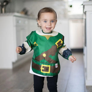 A toddler beams in a kitchen, wearing a Bumkins Sleeved Bib: Zelda™.