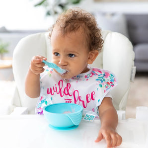 A toddler wearing a floral bib in a high chair enjoys a meal from a Bumkins Silicone First Feeding Set: Blue.