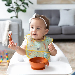 A baby in a high chair, with a white bow, eats from a Bumkins Silicone First Feeding Set: Clay in a cozy living room.