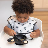 A toddler in a high chair enjoys bananas with a Bumkins Mickey Mouse Classic Black silicone spoon from the feeding set.