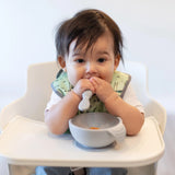A brown-haired baby sits in a high chair, wears a green bib, and holds Bumkins Taffy Silicone Dipping Spoons with a bowl in front.
