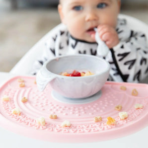 A baby grips a Bumkins Silicone First Feeding Set spoon, sitting among snacks on the Marble mat.