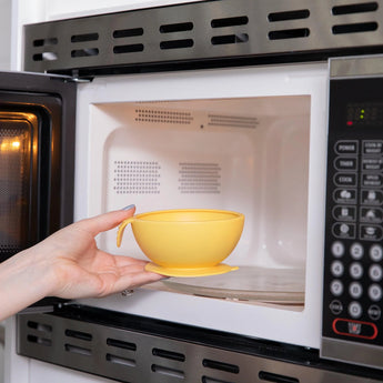 A hand places a Bumkins Silicone First Feeding Set: Pineapple into an open microwave.