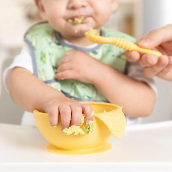 A baby with a green bib self-feeds, grabbing rice from a Bumkins Silicone First Feeding Set in Pineapple as a hand holds a spoon nearby.