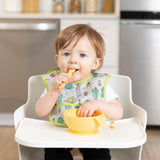 A toddler in a high chair uses a carrot stick from their Bumkins Silicone First Feeding Set: Pineapple, with a bib of playful designs.