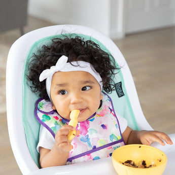 A baby with curly hair sits in a high chair wearing a colorful bib, ready to self-feed with Bumkins Pineapple Silicone Feeding Set nearby.