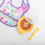 A vibrant baby bib featuring a floral design is spread next to a Bumkins Silicone First Feeding Set in Pineapple, which includes a yellow suction base bowl topped with food chunks. Nearby lies a matching yellow textured-handle spoon, ideal for promoting self-feeding.