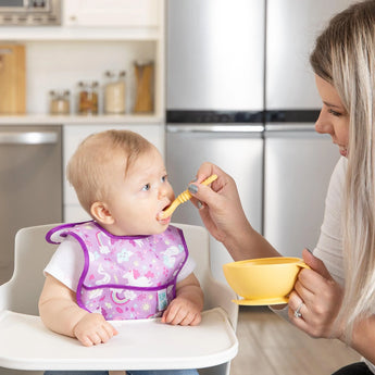 A woman feeds a baby in a high chair using a yellow spoon from the Bumkins Silicone First Feeding Set: Pineapple. The baby wears a purple bib.