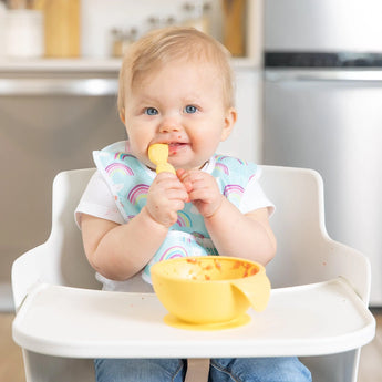 A baby self-feeds with a spoon and colorful bib, using Bumkins Silicone First Feeding Set: Pineapple, on a high chair tray.