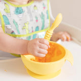A baby in a high chair self-feeds with a spoon from the Bumkins Silicone First Feeding Set: Pineapple while wearing a cactus-themed bib.