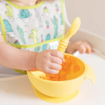 A baby in a high chair self-feeds with a spoon from the Bumkins Silicone First Feeding Set: Pineapple while wearing a cactus-themed bib.