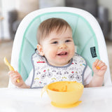 A happy baby in a high chair holds a spoon, ready for adventure with Bumkins Silicone First Feeding Set: Pineapple.