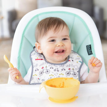 A happy baby in a high chair holds a spoon, ready for adventure with Bumkins Silicone First Feeding Set: Pineapple.