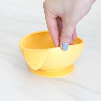 A hand with blue nail polish holds a yellow Silicone First Feeding Set: Pineapple by Bumkins, on a white surface.