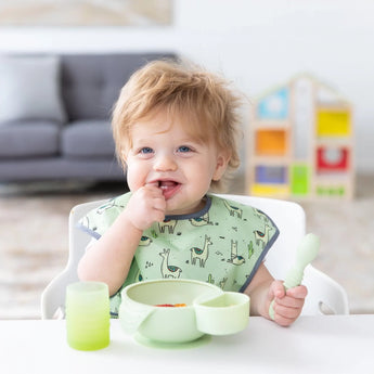 A toddler in a dinosaur bib enjoys mealtime with Bumkins Silicone First Feeding Set: Sage, surrounded by toys in soft focus.