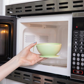 A hand places a Bumkins Silicone First Feeding Set: Sage bowl into an open microwave oven.