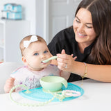 A woman feeds a baby with Bumkins Silicone First Feeding Set in Sage. The baby, in a floral bib and white bow, has a suction bowl and teether.