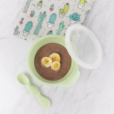 A bowl of chocolate pudding topped with bananas on marble, paired with Bumkins Silicone First Feeding Set in Sage and a cactus napkin.