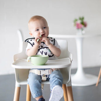 A baby in a white high chair enjoys mealtime with Bumkins Silicone First Feeding Set: Sage, a suction bowl and matching spoon.
