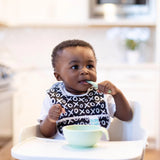 A toddler in a high chair eats using the Bumkins Silicone First Feeding Set in Sage, wearing a black and white X/O bib.
