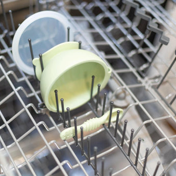The Bumkins Silicone First Feeding Set in Sage, with a suction bowl, lid, and spoon, can be placed on the top rack of the dishwasher.
