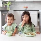 Two kids use their Bumkins Silicone First Feeding Set: Sage at the table, wearing matching green bibs with animal prints.