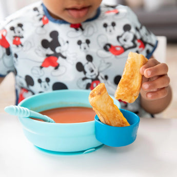 A child wearing a Mickey Mouse bib enjoys soup and breadsticks from a Bumkins Silicone Grip Bowl: Blue at a white table.