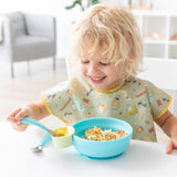 A smiling blonde child eats from Bumkins Silicone Grip Bowl: Blue at a white table, wearing a colorful bib and holding a spoon.