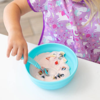 A child eats yogurt and fruit from Bumkins Silicone Grip Bowl: Blue, wearing a purple unicorn bib at a white table.