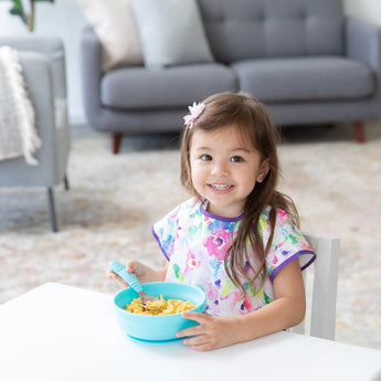 A cheerful child in a floral bib sits at a white table, eating macaroni and cheese from Bumkins Silicone Grip Bowl: Blue with a spoon.
