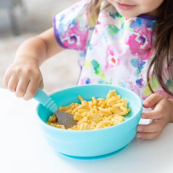 A toddler eats mac and cheese from Bumkins Silicone Grip Bowl: Blue at a white table, while wearing a vibrant bib.