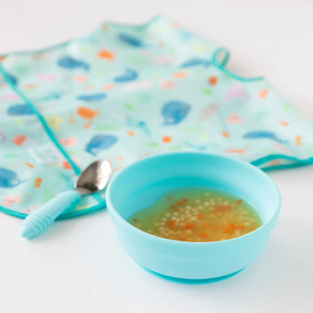 Bumkins Silicone Grip Bowl: Blue with soup and pasta, a blue spoon, and ocean-themed bib on a white background.