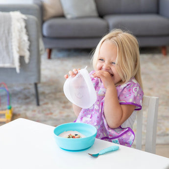 A child with long blonde hair in a purple bib holds a transparent lid over a Bumkins Silicone Grip Bowl: Blue filled with yogurt on a table.