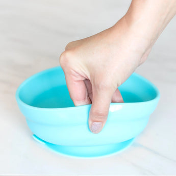 A hand holds a Bumkins Silicone Grip Bowl: Blue, demonstrating the suction cup bottom on a marble surface.
