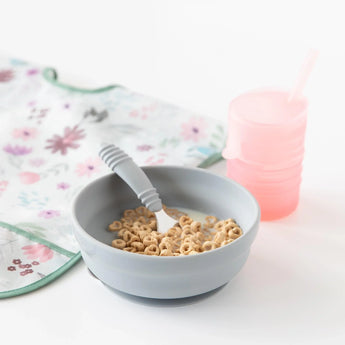 Bumkins Silicone Grip Bowl: Gray with cereal and milk, a gray spoon, a pink sippy cup, and floral bib on a white background.