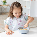 A young girl uses a spoon to eat cereal from Bumkins Silicone Grip Bowl: Gray, wearing a floral bib at a white table.