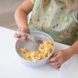 A child enjoys mac and cheese with a fork from Bumkins Silicone Grip Bowl: Marble at a white table while wearing a colorful bib.