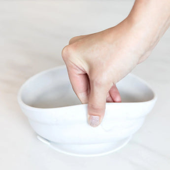 A hand holds a Bumkins Silicone Grip Bowl: Marble, demonstrating the suction cup bottom on a marble surface.