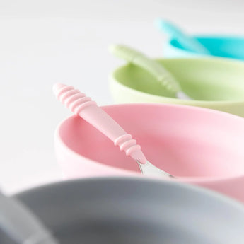 Close-up of four bowls and spoons, including Bumkins Silicone Grip Bowl: Pink, on a white background.