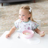 A happy toddler in a floral bib enjoys ice cream from Bumkins Silicone Grip Bowl: Pink at a white table, paired with a cookie.