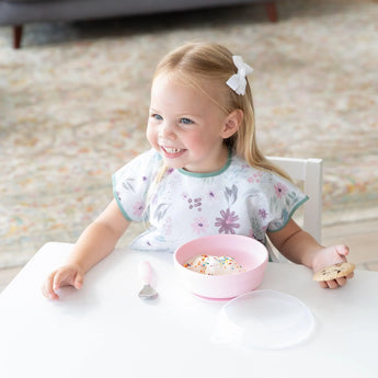 A happy toddler in a floral bib enjoys ice cream from Bumkins Silicone Grip Bowl: Pink at a white table, paired with a cookie.