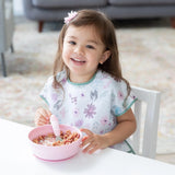 A smiling toddler with brown hair enjoys cereal from Bumkins Silicone Grip Bowl: Pink at a table, wearing a floral bib and using a spoon.