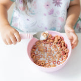 A child wearing a floral bib enjoys colorful cereal and milk from Bumkins Silicone Grip Bowl: Pink at a white table, using a spoon.