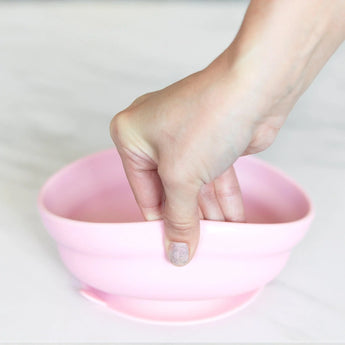 A hand holds a Bumkins Silicone Grip Bowl: Pink, demonstrating the suction cup bottom on a marble surface.