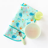 Bumkins Silicone Grip Bowl: Sage sits on a white background next to a folded ocean-themed bib, a green spoon, and a green cup.