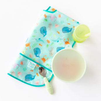 Bumkins Silicone Grip Bowl: Sage sits on a white background next to a folded ocean-themed bib, a green spoon, and a green cup.